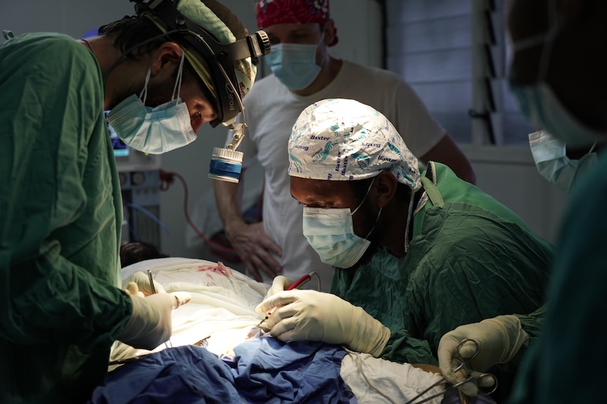 Three doctors wearing surgical masks, caps, gowns and gloves operate on a patient
