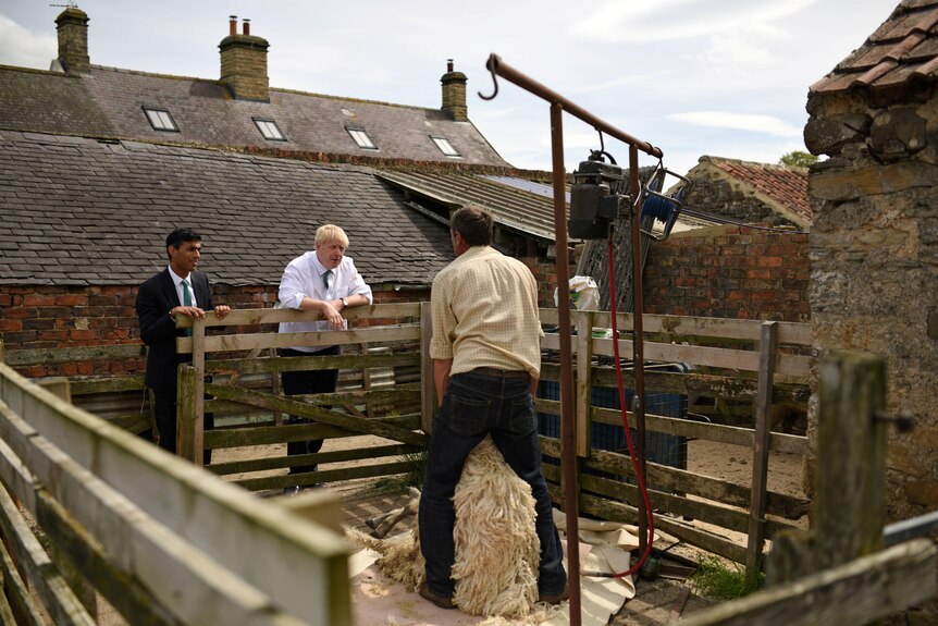 Ο Μπόρις Τζόνσον και ο Ρίσι Σουνάκ φορώντας κοστούμια συνομιλούν με έναν άντρα που κρατά ψηλά ένα πολύ χνουδωτό πρόβατο