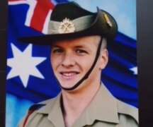 A man wearing an army uniform and army hat standing in front of the Australian flag. 