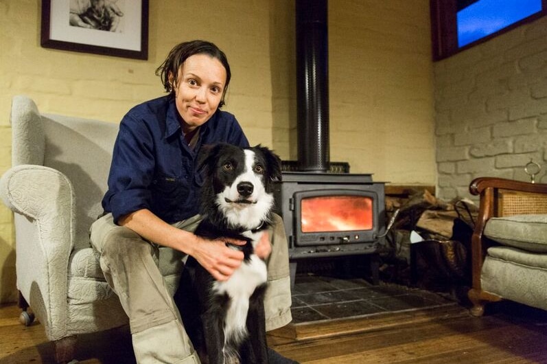 Trainer Fiona Jackson sitting on a chair with her border collie dog sitting between her legs in a lounge room with a fireplace