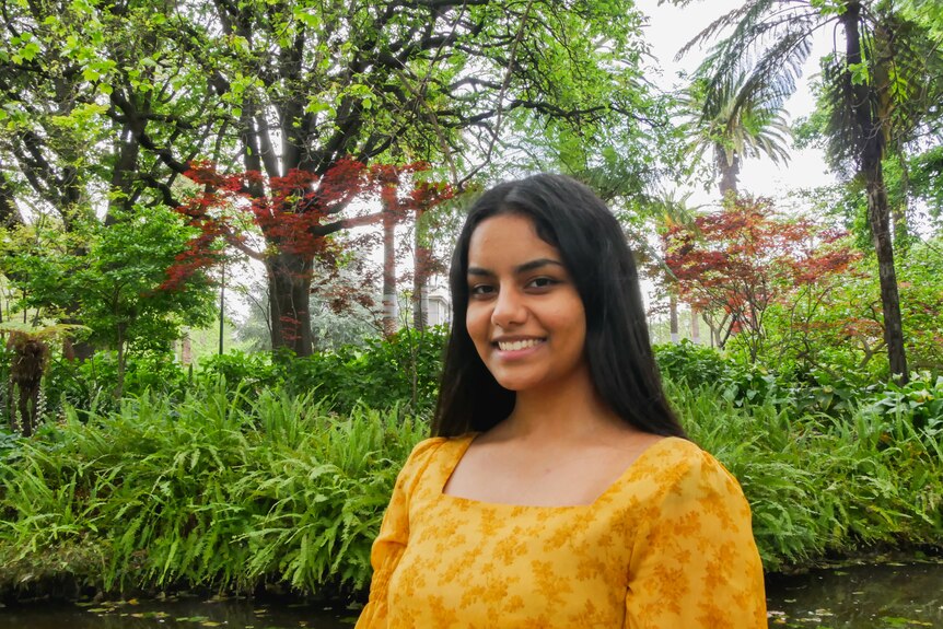 Tanya wears a yellow top and looks to the camera with a smile while standing in front of a lake with trees behind.