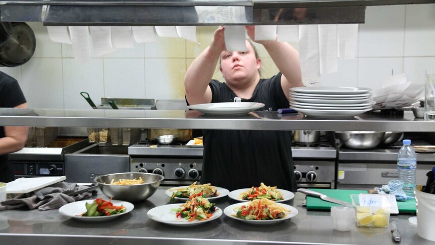 A female chef stands in a kitchen reaching up to look at a line of meal orders above plates prepared with salad and chips.