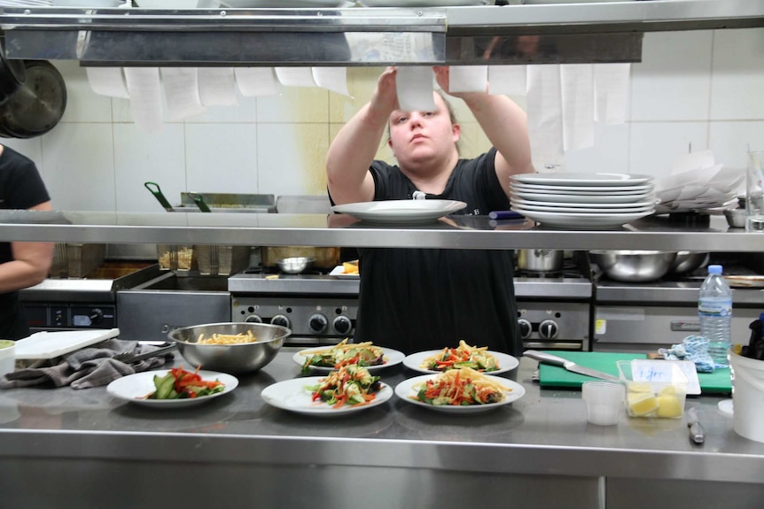 A female chef stands in a kitchen reaching up to look at a line of meal orders above plates prepared with salad and chips.