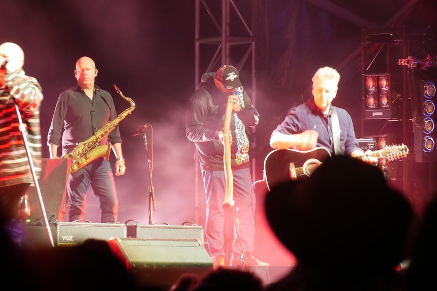 A man playing a didgeridoo faced down with a man on the right with a guitar, a man on the left with a saxophone.
