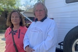 An older man in a white hoodie and woman in a melon-coloured hoodie stand in front of a caravan.