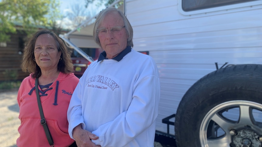 An older man in a white hoodie and woman in a melon-coloured hoodie stand in front of a caravan.