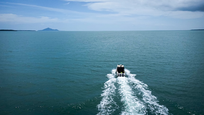 An aerial view from behind a speed boat shows it heading out on a clear day towards two land masses on the horizon