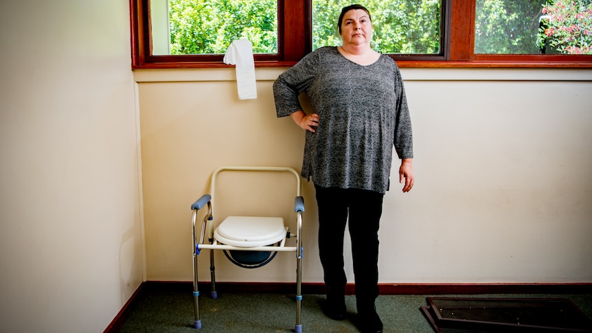 Shelly stands with her hand on her hip, next to a portable toilet inside her home.