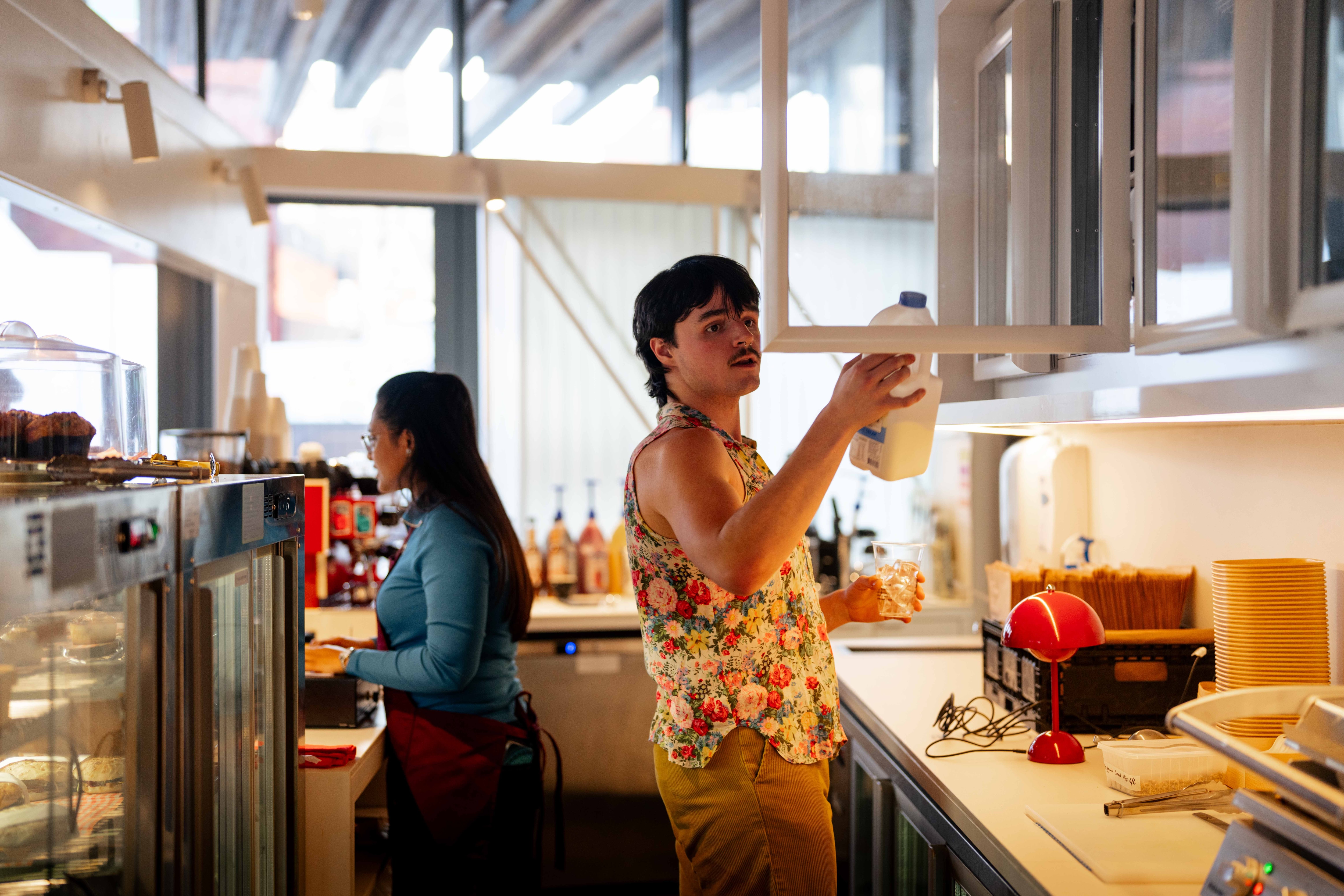A woman takes someone's order whilst a man puts milk in a cupboard