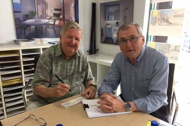 two men at a table in a real estate office smile as they sign a cheque