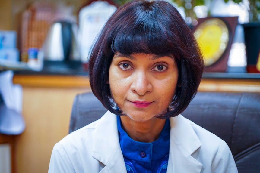 An Indian doctor with dark chin-length hair and a lab coat sitting at a desk
