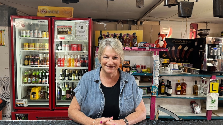 A lady in a blue shirt and grey hair stands smiling leaning on a bar.
