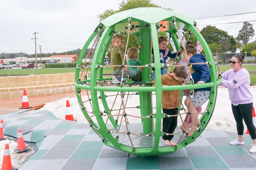 A group of children in a large green wheel at a playground with a mum pushing it