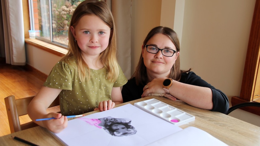 A woman kneels next to a young child drawing on a table