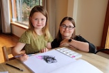 A woman kneels next to a young child drawing on a table