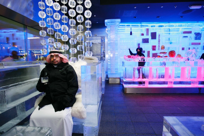 A man in a dishdashi sitting in an ice bar with a drink in his gloved hand