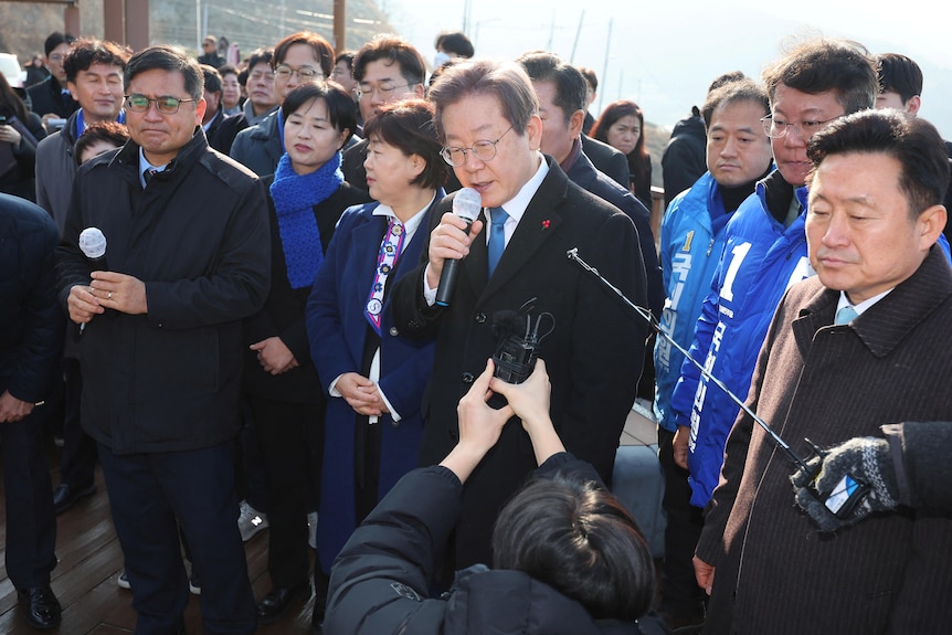 A man is speaking into a microphone with a crowd surrounding him.