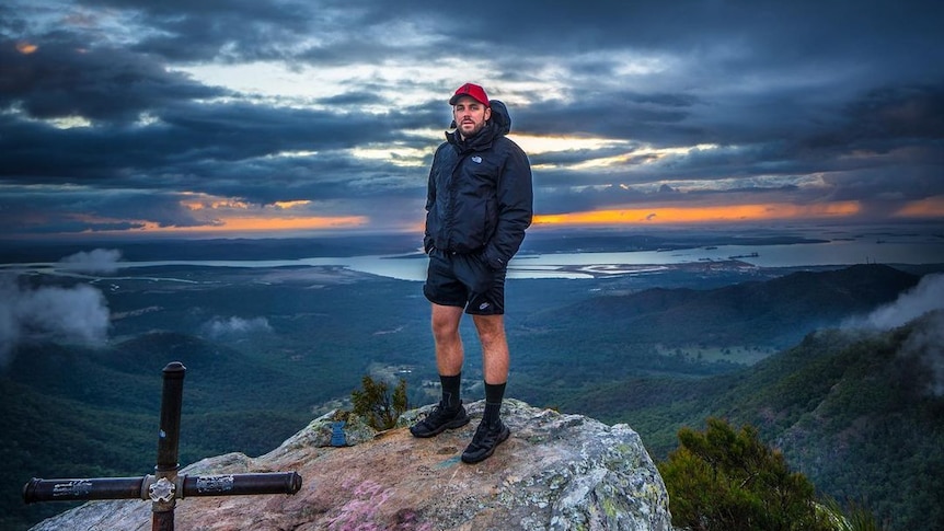A man in a jumper, shorts and shoes standing on a mount top, ocean, trees, clouds behind, dark eerie colours.