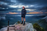 A man in a jumper, shorts and shoes standing on a mount top, ocean, trees, clouds behind, dark eerie colours.