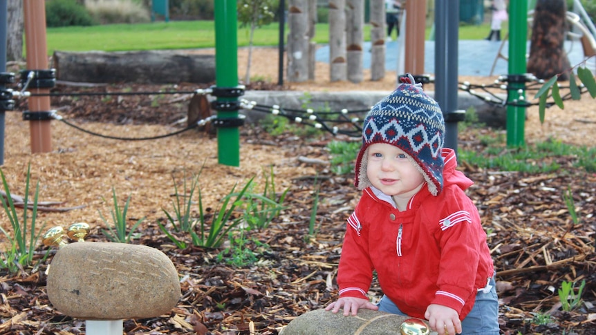 A 10 month old boy in a beanie plays with a metal bell on a rock, playground in background.