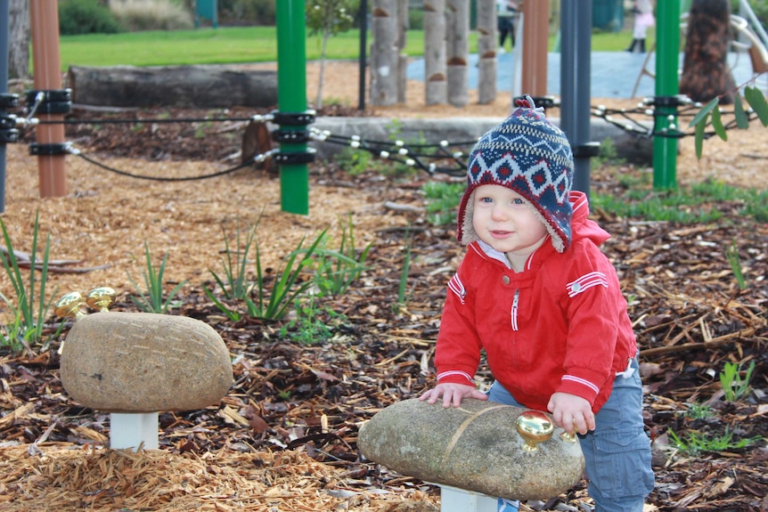 A 10 month old boy in a beanie plays with a metal bell on a rock, playground in background.