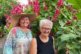 Two women in gardening clothes and hats in front of a large bush with hot pink flowers.