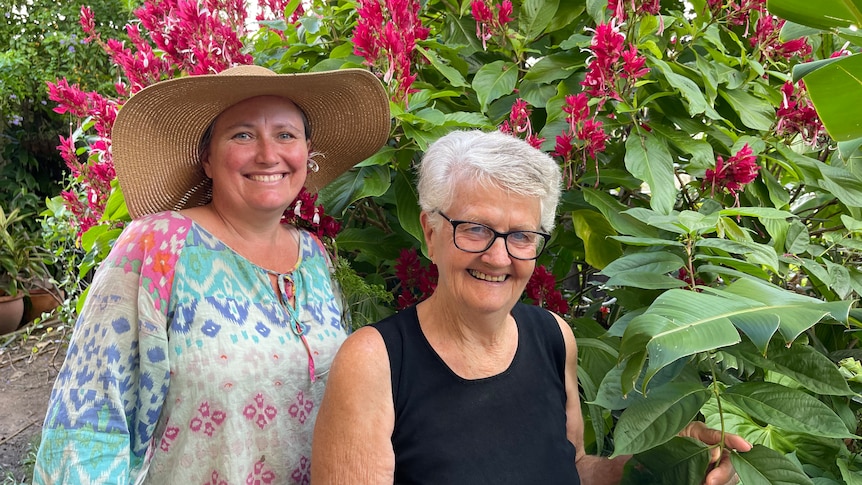 Two women in gardening clothes and hats in front of a large bush with hot pink flowers.