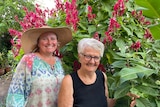 Two women in gardening clothes and hats in front of a large bush with hot pink flowers.
