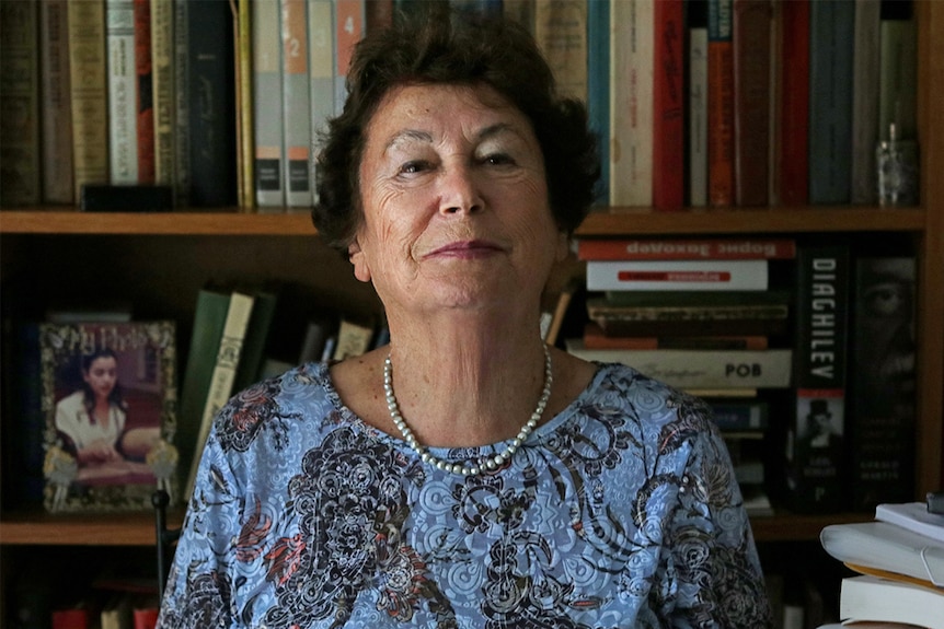 An older woman stands in front of a bookshelf, chin raised, smiling at the camera.