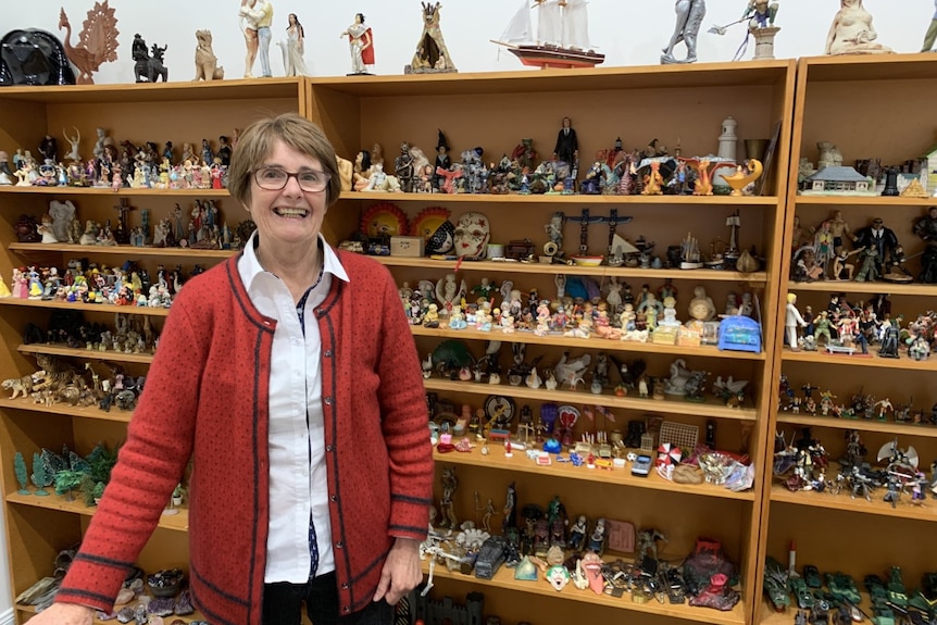 Woman in a red cardigan and glasses smiles at the camera. There are shelves of assorted knick-knacks behind her.