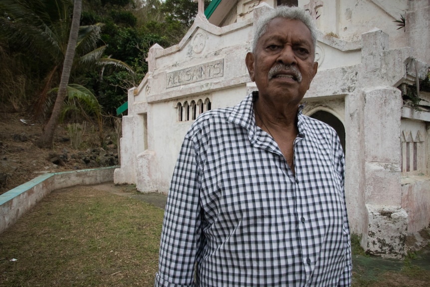 An older man stands outside the front of a crumbling stone church that is painted white with a sign that reads 'All Saints'.