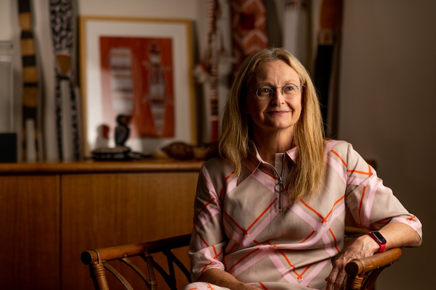 A woman with long blonde hair and glasses sitting on a chair inside a darkened office and smiling.