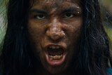 A sooty and muddy teen boy with long hair and nose and eyebrow piercing speaks with snarling and serious expression in jungle.