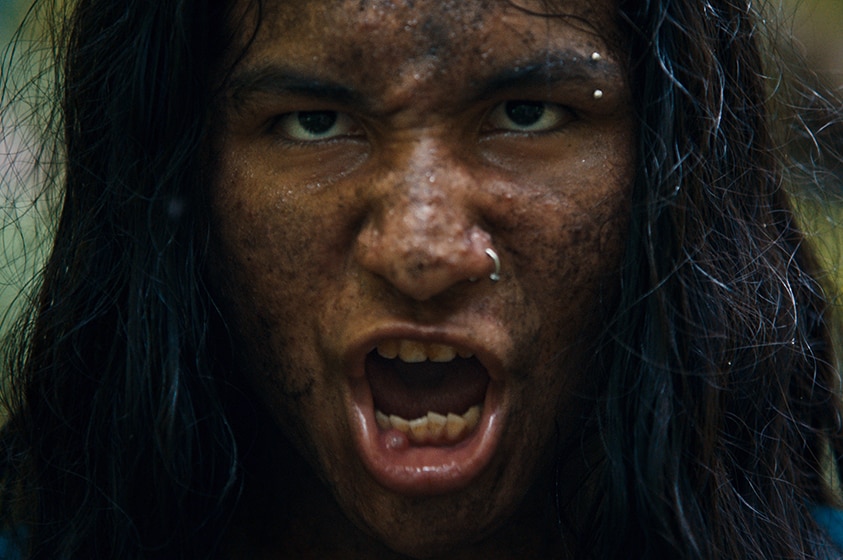 A sooty and muddy teen boy with long hair and nose and eyebrow piercing speaks with snarling and serious expression in jungle.