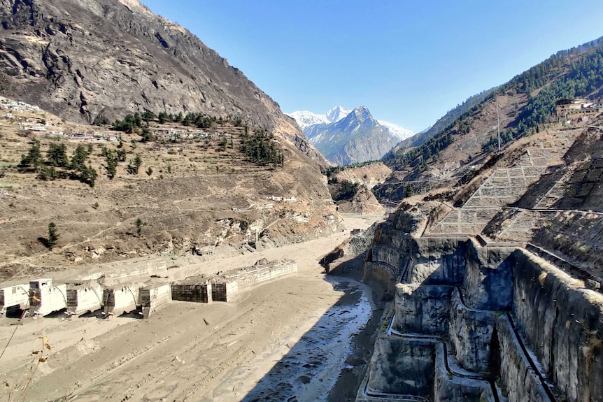 An aerial view shows a partially buried hydroelectric plant sitting in a deep valley with snowy peaks in the far background.