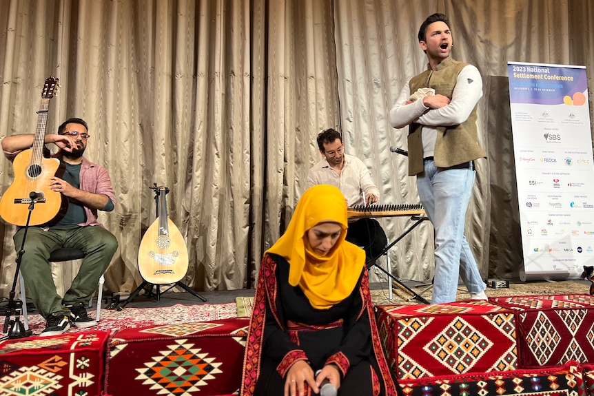 a woman in yellow hijab, black dress sitting in front of the stage, with three man on the stage behind her.