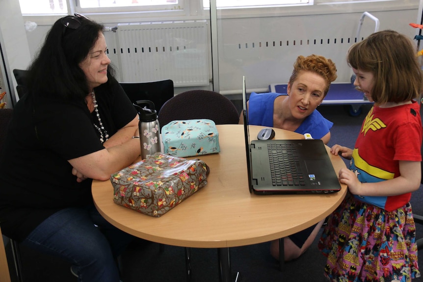 Two women and a young girl gather around a laptop.