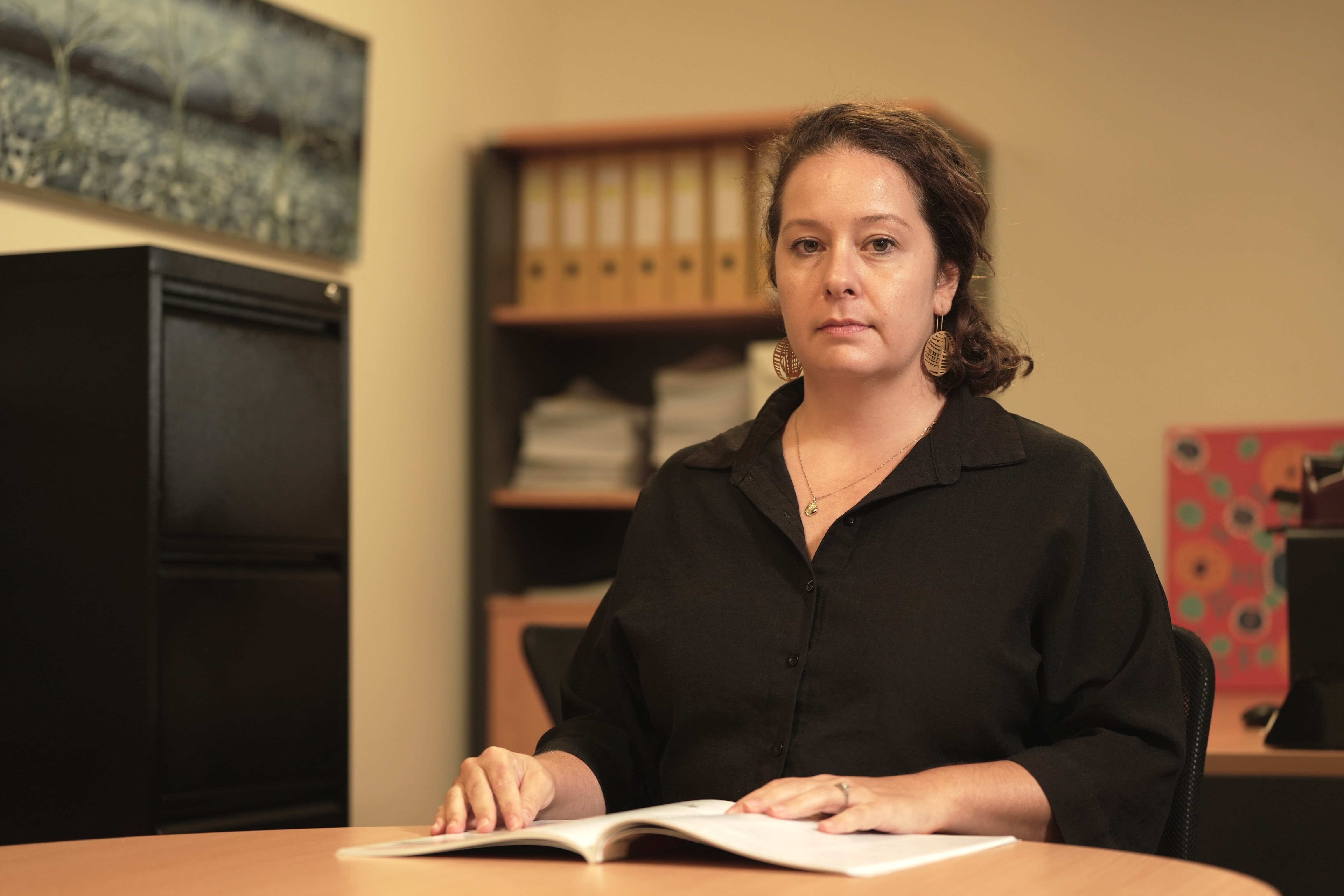 A white woman with brown hair, wearing a black button-up shirt, sitting in an office space with a report open on her desk.