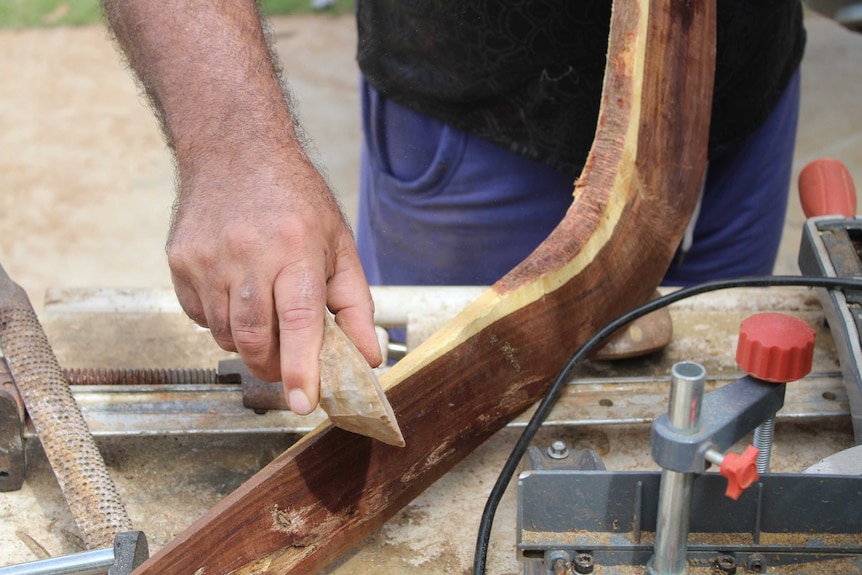 A man uses a stone tool to craft a traditional boomerang