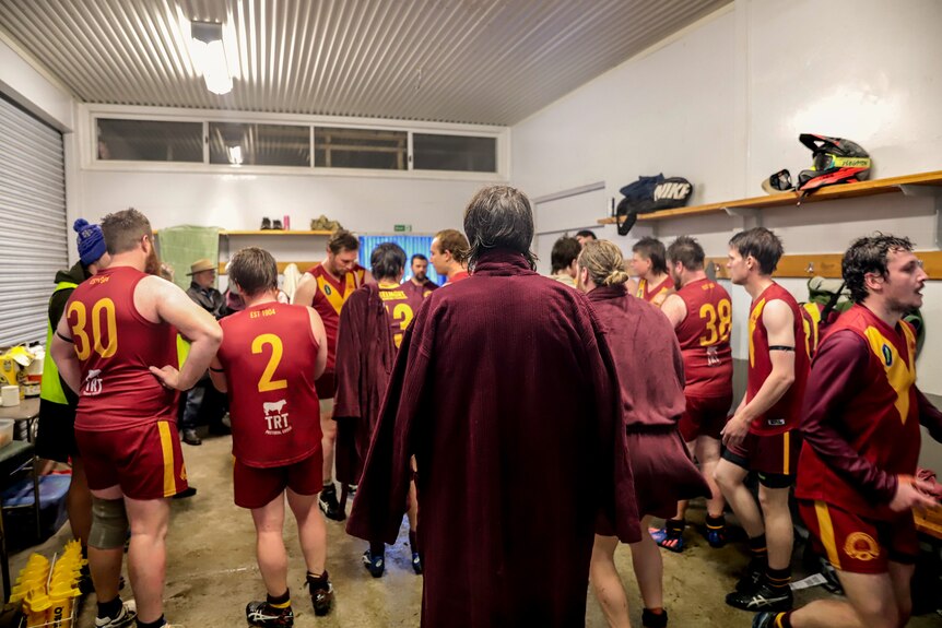A man stands in a maroon robe stands amid other football players in a fluorescent lit change room