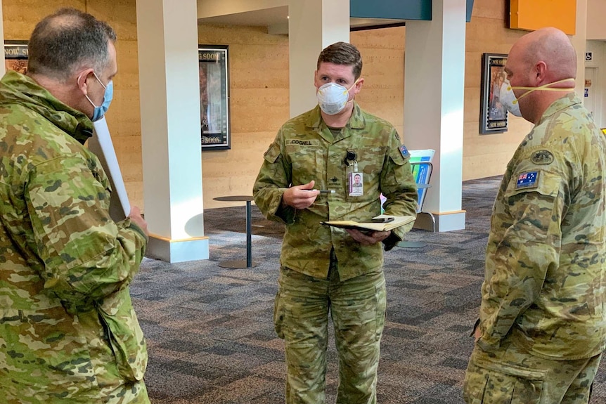 Three defence force members wearing military uniform and masks stand together talking.