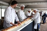 Four bakers hold different parts of a really long baguette entering a rotating oven.