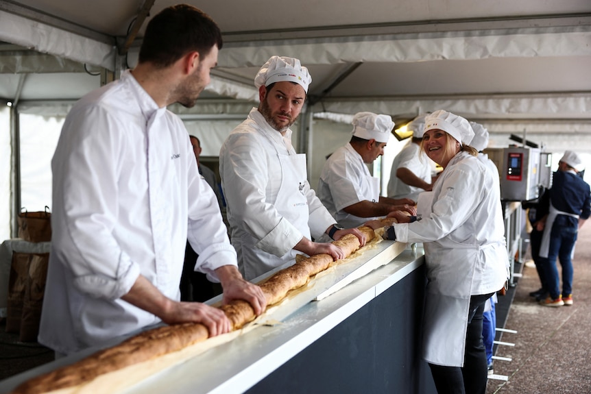 Four bakers hold different parts of a really long baguette entering a rotating oven.