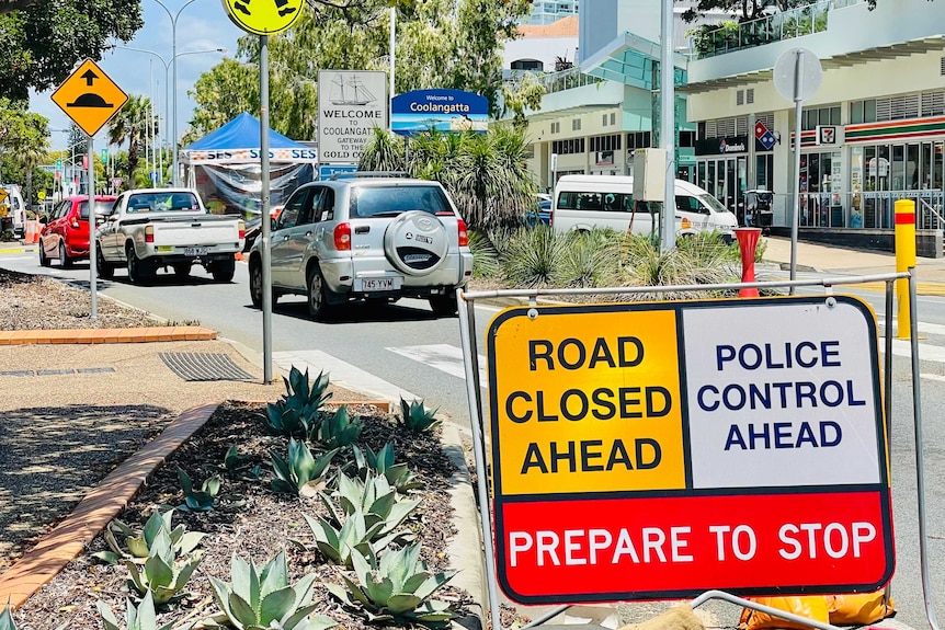 A road closed/police control sign on a narrow city street on a sunny day with trees in background.