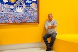 A man sitting alongside a colourful painting of Sydney Harbour.