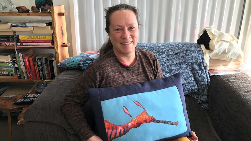 Benita Vincent, AKA Benny Marine, sitting on a couch holding a pillow with a sea-dragon illustration on it