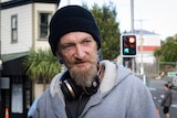 A man with a medium-length beard wearing a black beanie, headphones around his neck and a grey hoodie