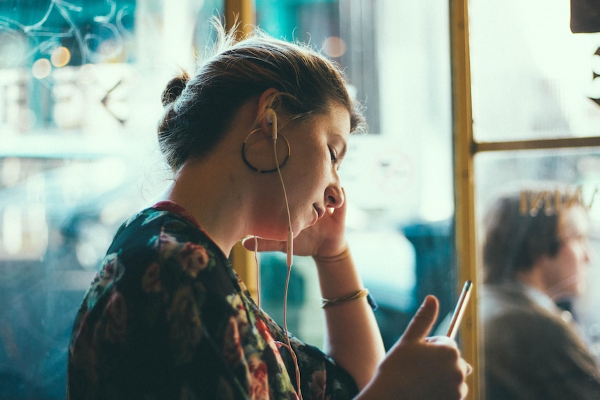 Woman sitting, leaning on window, wearing earbuds and looking at phone