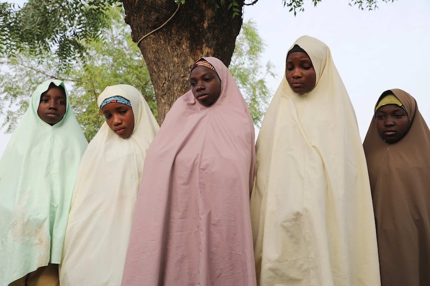Five girls wearing long head scarfs stand next to a tree