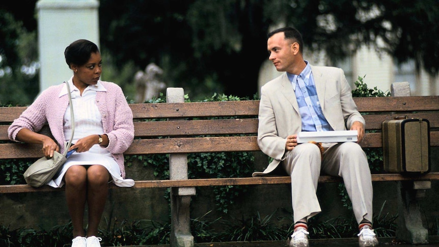 Forrest Gump sits on a park bench looking at the woman, who is looking back at him